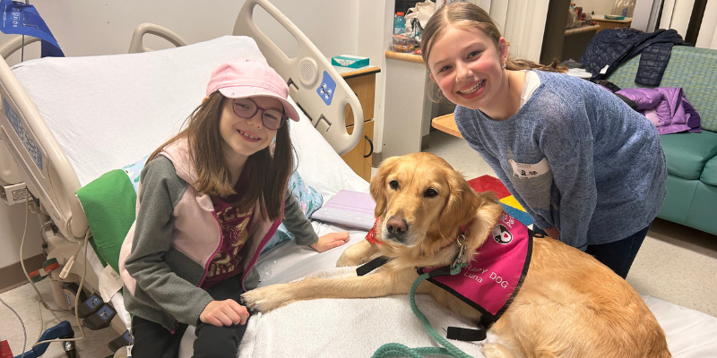 Zofia in a hospital bed with her sister and a therapy dog